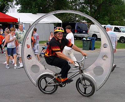 [Man on a bicycle wearing a helmet with flames and flames on his shirt and pants sitting inside a large wheel.]